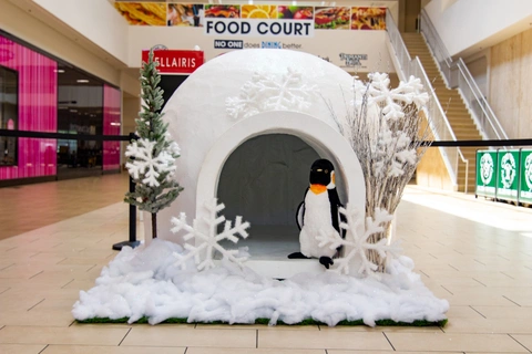 A mall display with an igloo surrounded by faux snow and white trees. A plush penguin stands at the entrance. Bright, festive atmosphere.