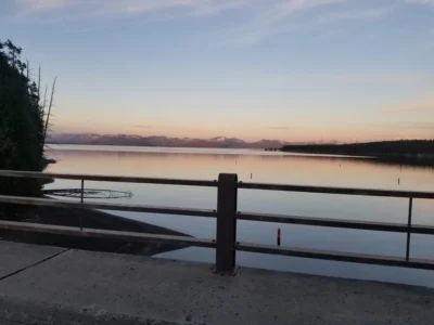 Calm lake at sunset with mountains in the distance, partially framed by a bridge railing. The sky is softly colored, creating a serene atmosphere.