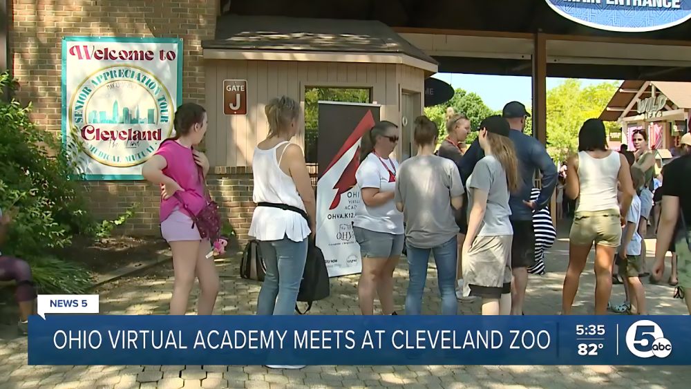 A group of people gather near a gate at Cleveland Zoo. A "Welcome to Cleveland" sign is visible. The atmosphere is casual and lively.