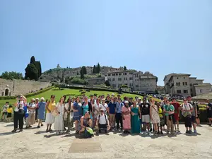 A large group of travelers posing together outdoors in front of a hillside town with stone buildings, green lawns, and tall cypress trees under a clear blue sky. The group stands closely together, smiling, with some people wearing hats, sunglasses, or backpacks.