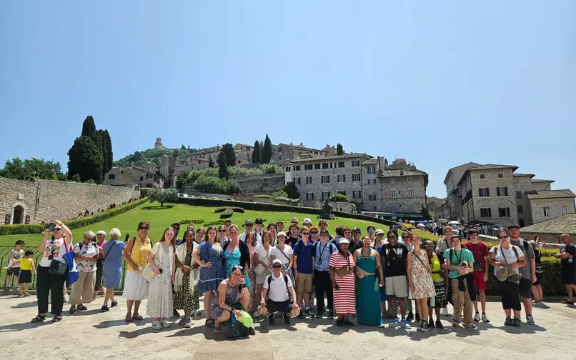 A large group of travelers posing together outdoors in front of a hillside town with stone buildings, green lawns, and tall cypress trees under a clear blue sky. The group stands closely together, smiling, with some people wearing hats, sunglasses, or backpacks.
