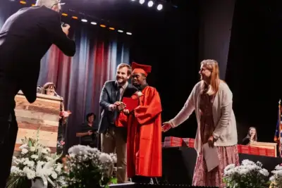 A graduate in a red cap and gown proudly holds a diploma, flanked by two smiling adults.