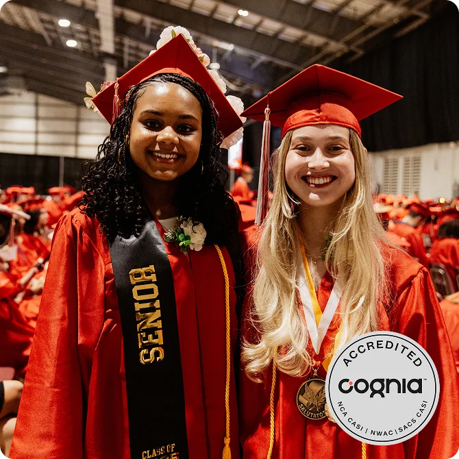 Two smiling graduates in red caps and gowns. One wears a "Senior" sash, the other a medal. Background shows fellow graduates seated. Jubilant mood.