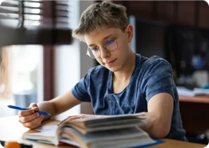 A young person with glasses studies at a desk, holding a pen and reading a book. The setting is calm and focused, with natural light from a window.