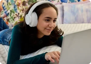A young woman with headphones smiles while using a laptop, lying on a bed with colorful pillows. She seems engaged and relaxed in a cozy setting.