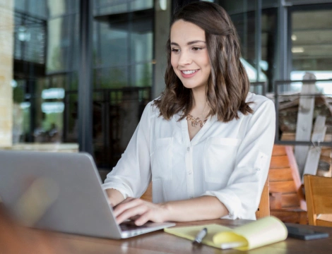 Smiling woman in a white blouse types on a laptop at a wooden table with a yellow notepad and pen. Modern glass office setting, focused and cheerful.