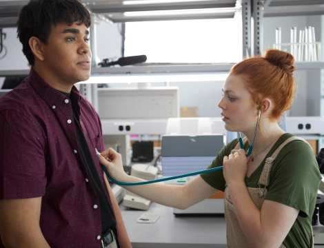 A woman uses a stethoscope to listen to a man's heartbeat in a lab setting. She wears overalls and focuses intently, conveying a professional tone.