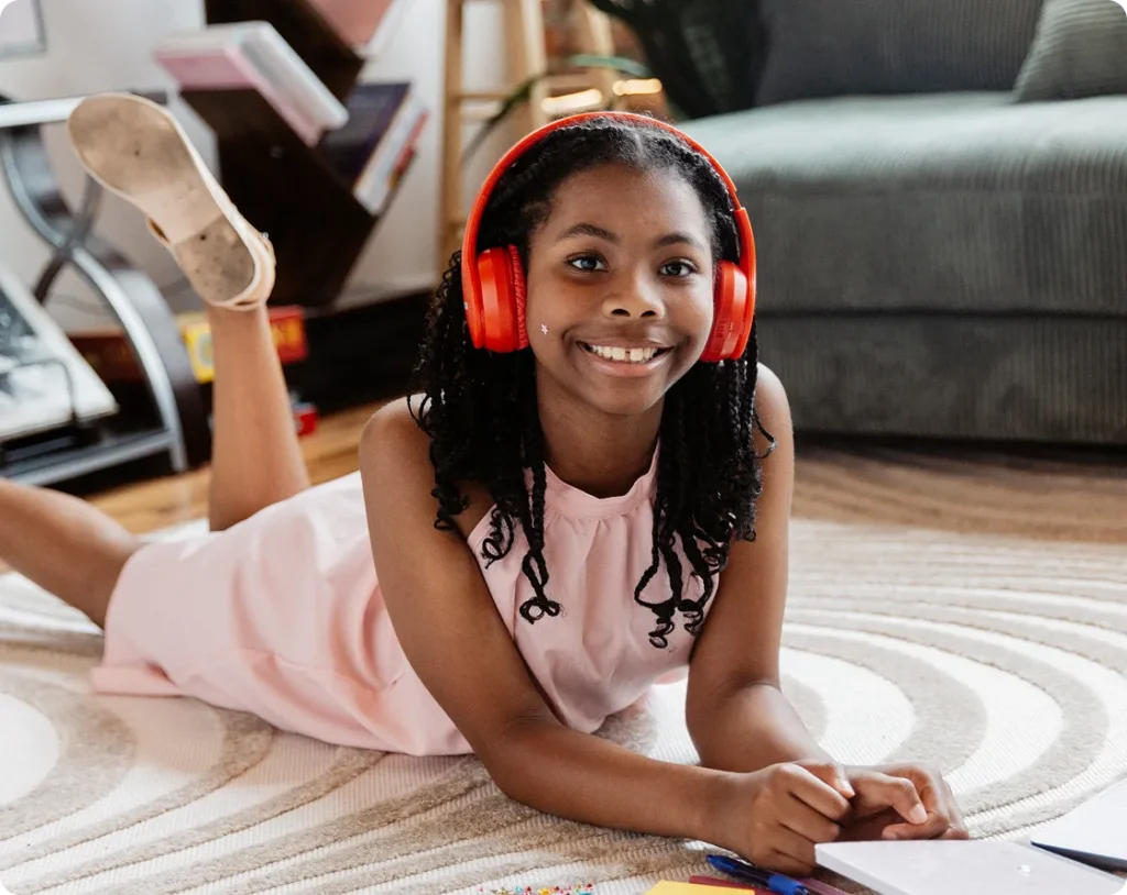 Young girl lying on a rug, smiling, wearing red headphones and a pink dress, surrounded by books and colored pencils. The scene is cozy and playful.