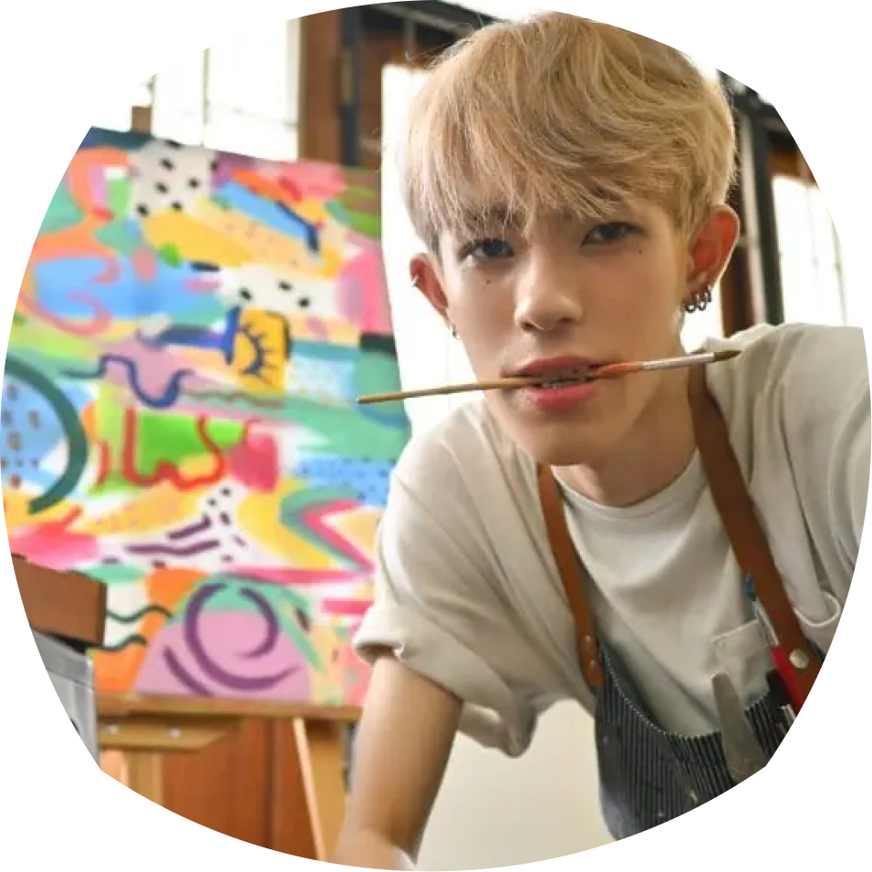 Young artist with a blond bob haircut, wearing a white shirt and apron, holds a paintbrush in his mouth. A colorful abstract painting is in the background.