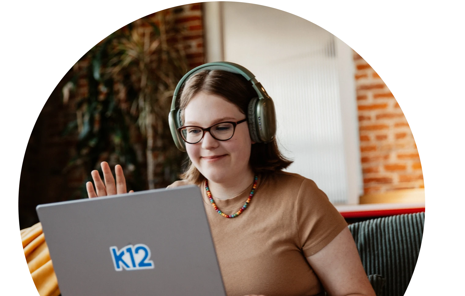 Young woman wearing headphones and glasses smiles while waving at a laptop. She wears a colorful necklace. Background shows a brick wall and plants.