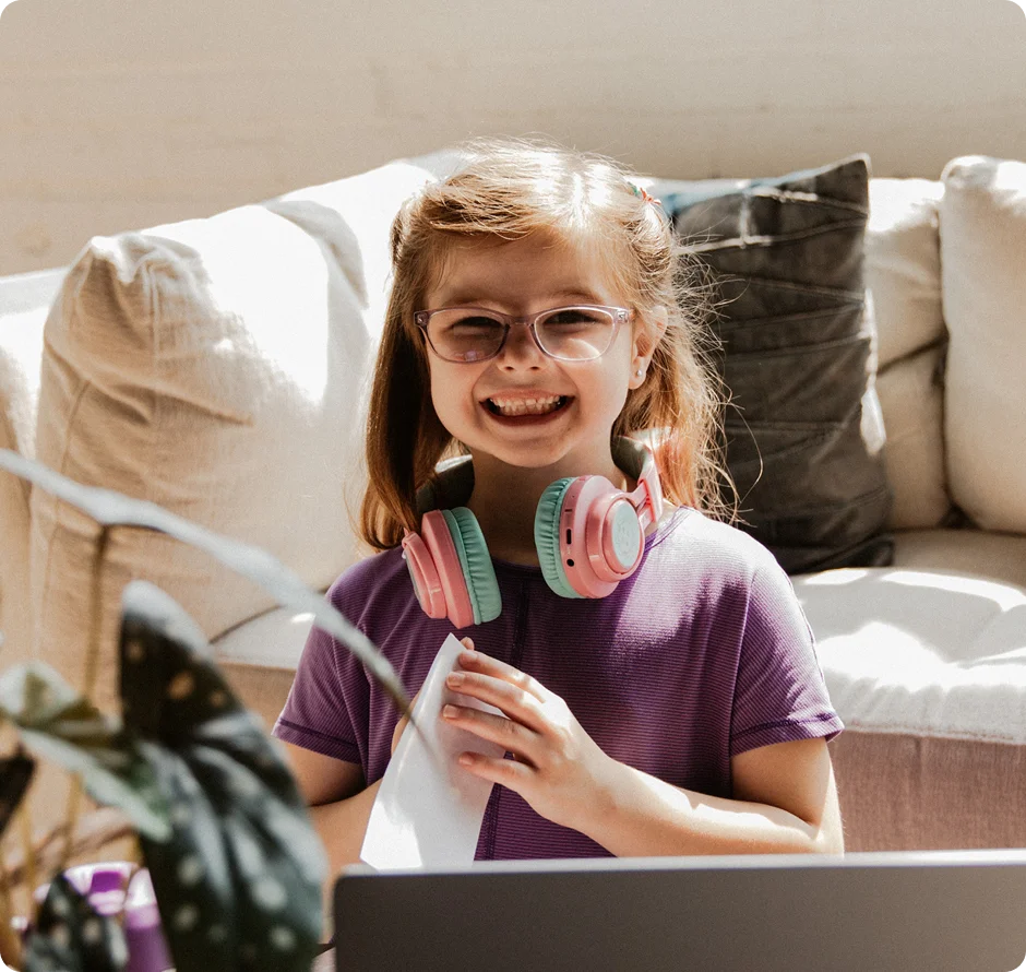 Smiling young girl with glasses and pink headphones sits on a couch, holding a paper. Natural light and a cozy ambiance fill the room.