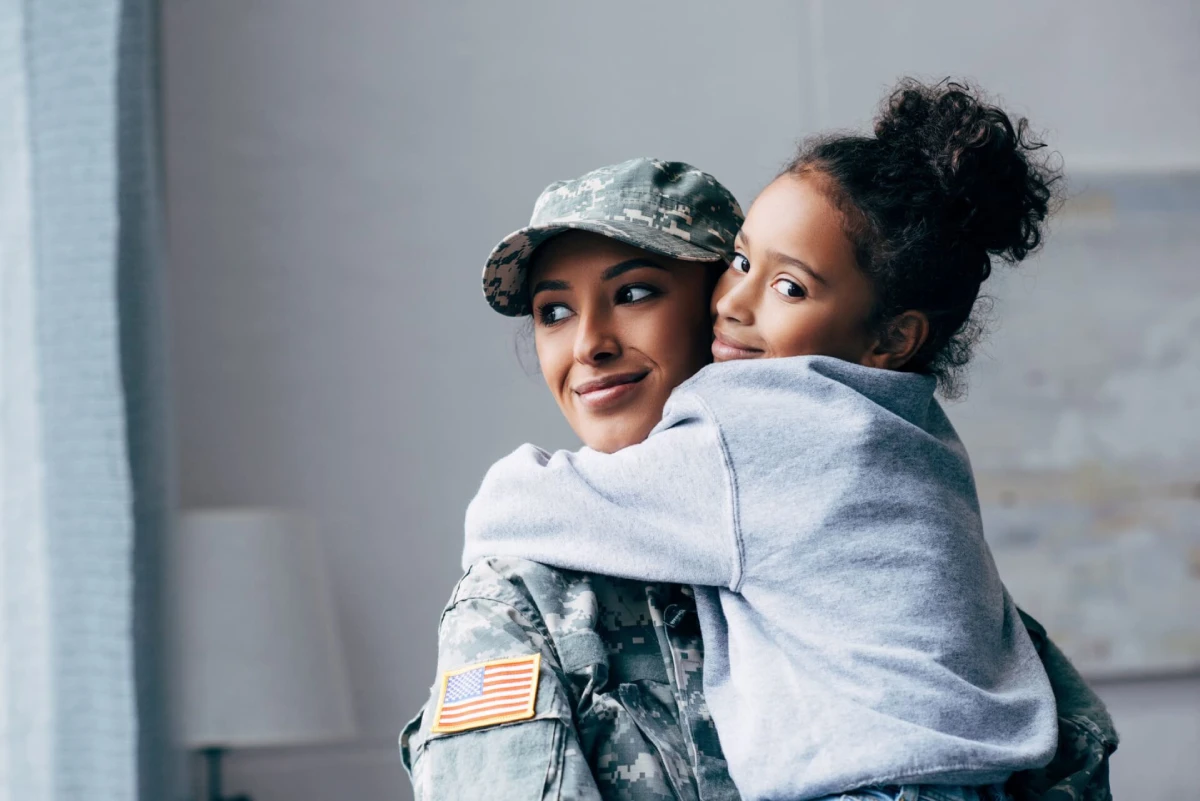 A woman in military uniform smiles gently as a young girl hugs her warmly, both exuding happiness and love. An American flag patch is visible on the uniform.