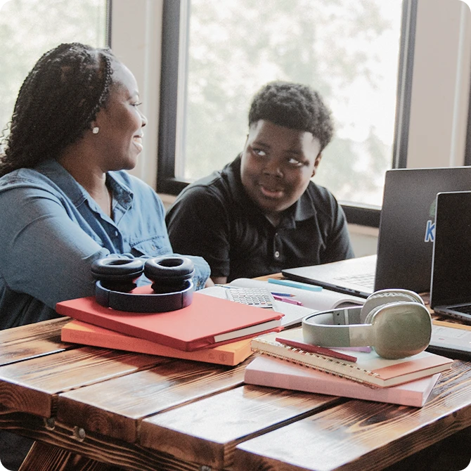 A woman and a child sit at a wooden table, smiling at each other beside laptops. Headphones and notebooks are scattered on the table, creating a warm, collaborative atmosphere.