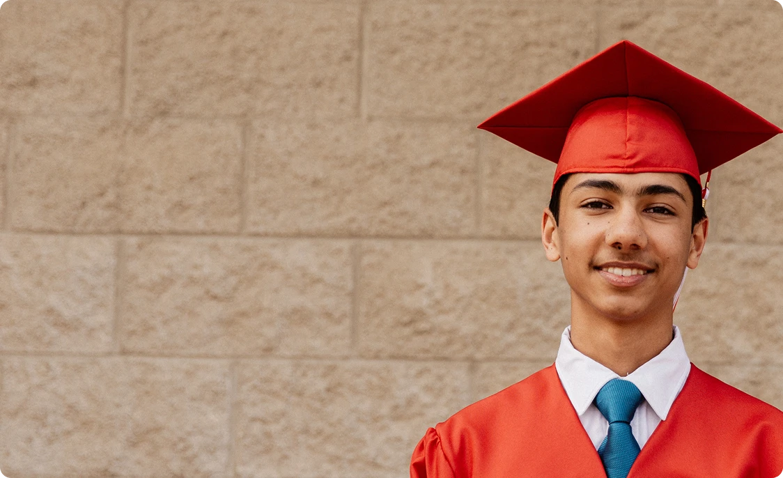 Smiling graduate in a red cap and gown stands against a light brick wall. He wears a white shirt and teal tie, exuding pride and accomplishment.