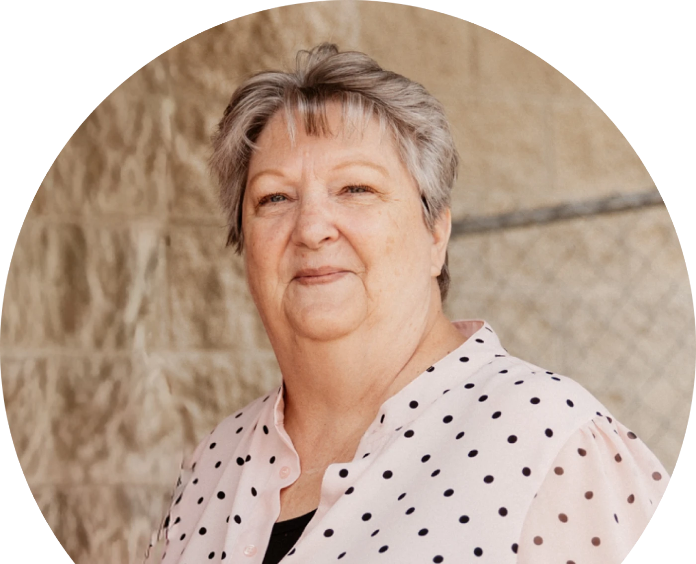 Elderly woman with short gray hair smiles softly wearing a white polka dot blouse. Background shows a textured, beige wall with a chain-link fence.