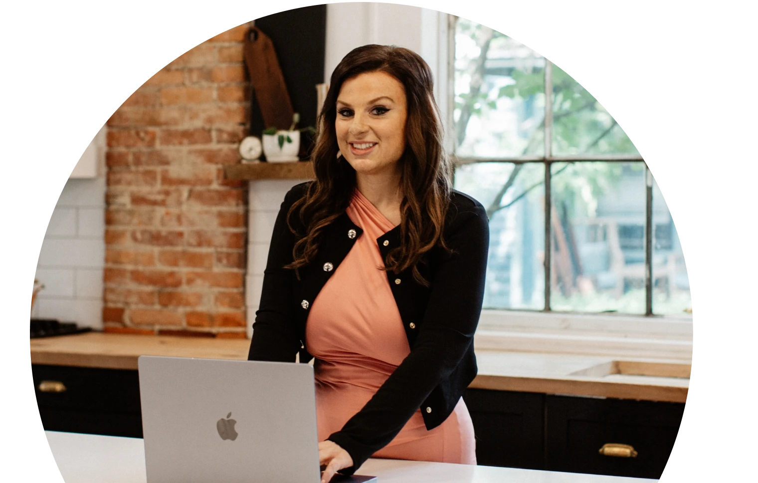 Woman in a pink dress and black cardigan stands at a kitchen counter with a laptop, smiling confidently. Background has brick walls and a large window.