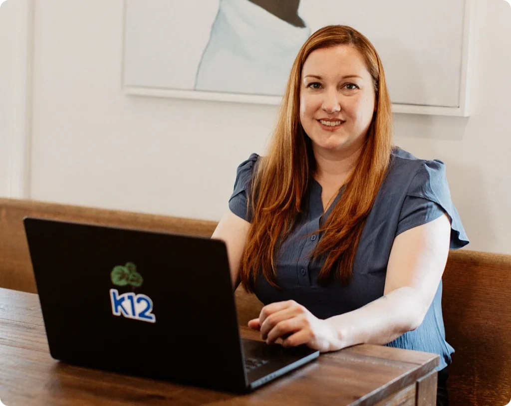 A woman with long red hair and a blue blouse smiles while sitting at a wooden table with a laptop displaying the "k12" logo, conveying a friendly and professional atmosphere.