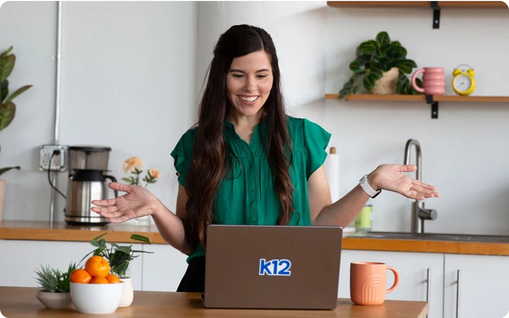 Smiling woman in green blouse gestures while video conferencing on a laptop with "k12" logo, set in a bright kitchen with plants and mugs.