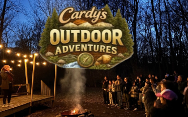 A group of people stands near a crackling campfire in a wooded area at dusk. Strings of lights are hung nearby. Above them is a large, stylized logo that reads "Cardy's Outdoor Adventures," featuring pine trees and mountains.