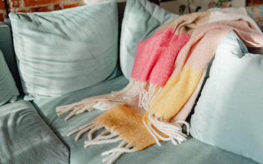 cozy corner club image A plush, light blue sofa with a colorful, fringed blanket draped over it. The blanket features stripes in shades of pink, yellow, and beige. In the background, part of a brick wall is visible.